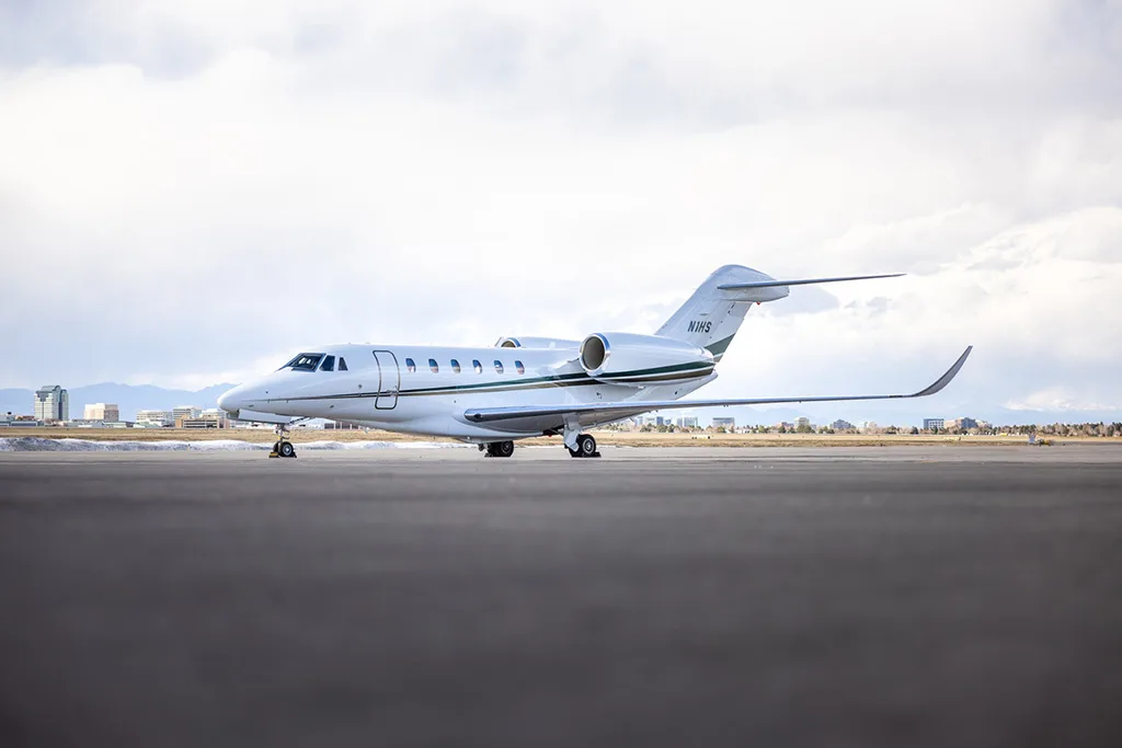 A white business jet with green and gold stripes on the tail, registration N1HS, parked on an airfield with a city skyline in the background.