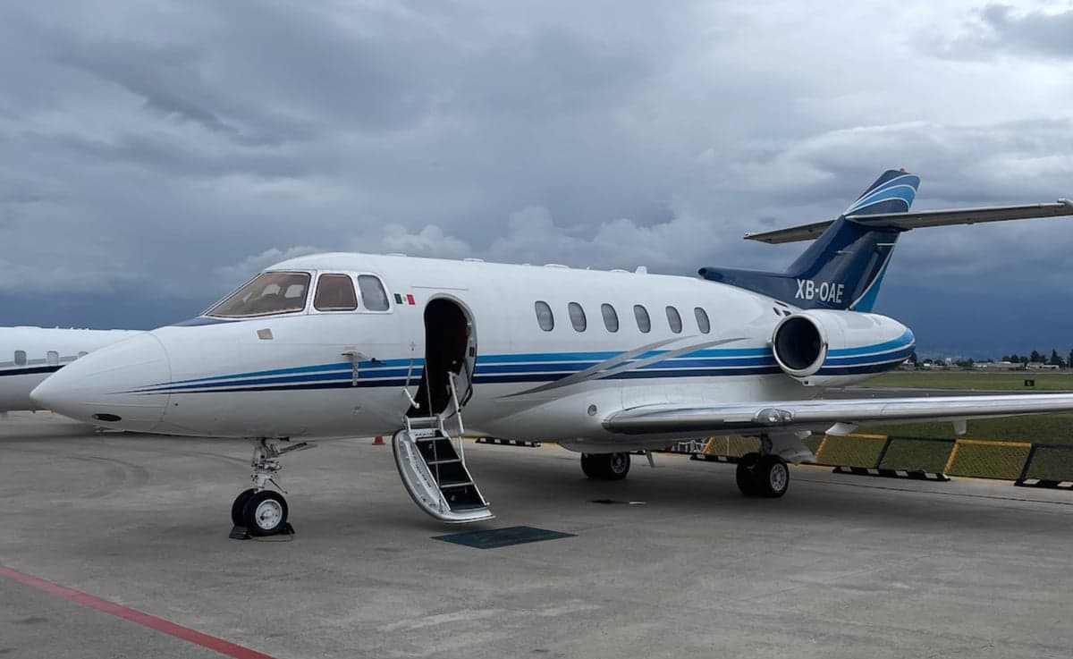 A white business jet with blue stripes (XB-OAE) parked on a tarmac with its passenger door open and stairs extended.