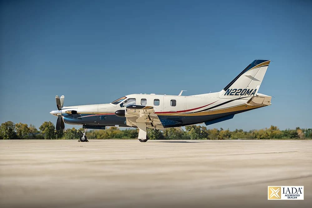 A white and blue turboprop aircraft with gold and red stripes, registration N220MA, parked on a tarmac under a clear blue sky.