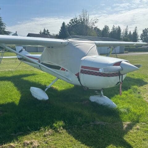 A Cessna 150 aircraft with a white and black paint scheme and red accents, covered with a gray cover, parked on the grass.