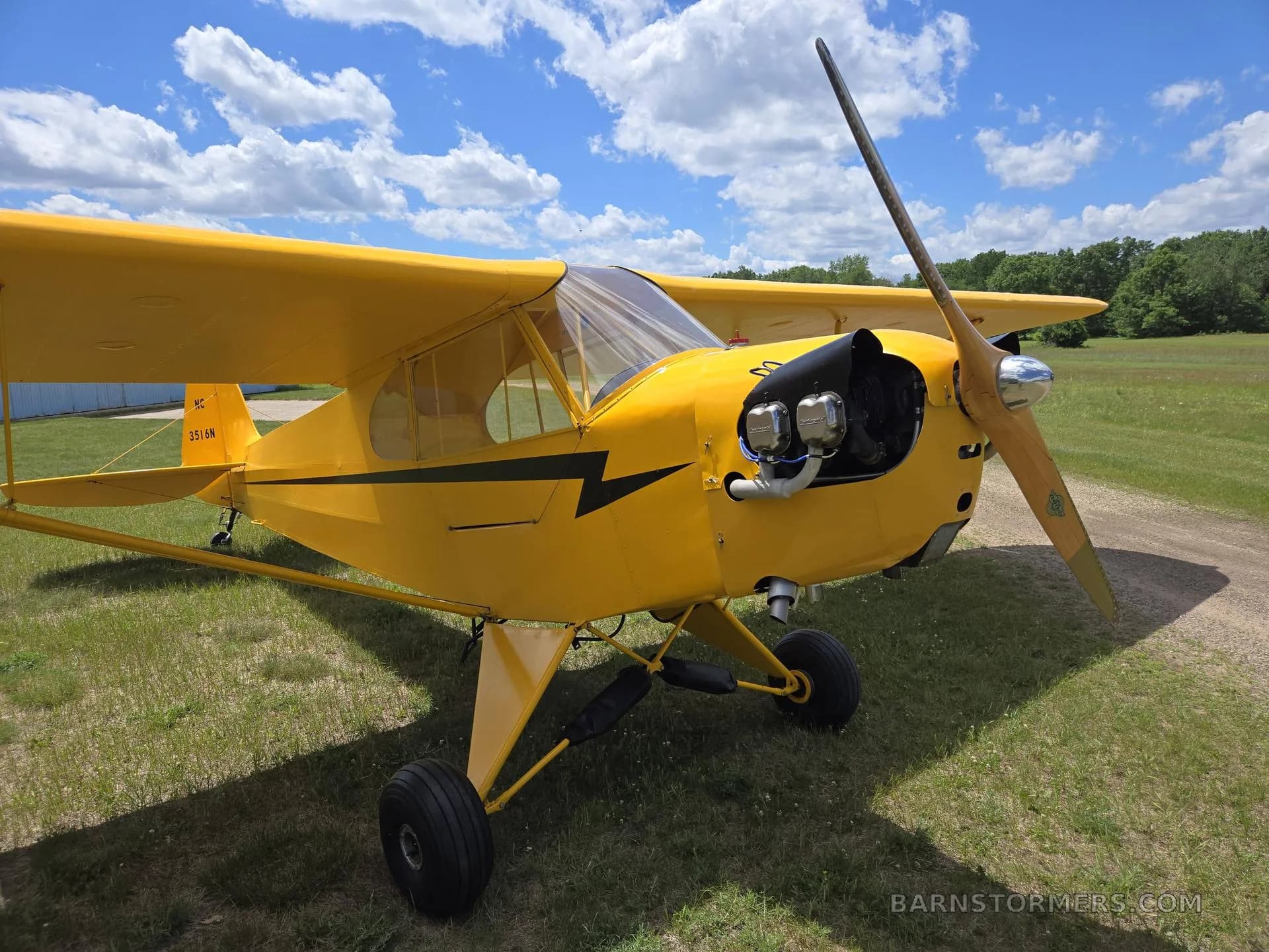 A close-up front view of a yellow Piper Cub-style aircraft with its engine exposed and a wooden propeller.