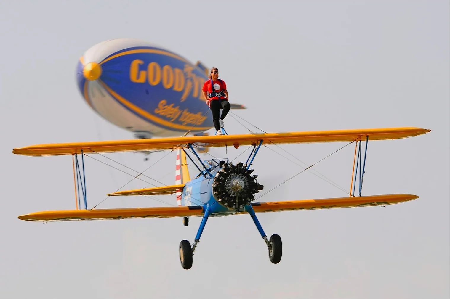 A biplane with a person standing on the wing flies in front of a Goodyear blimp.