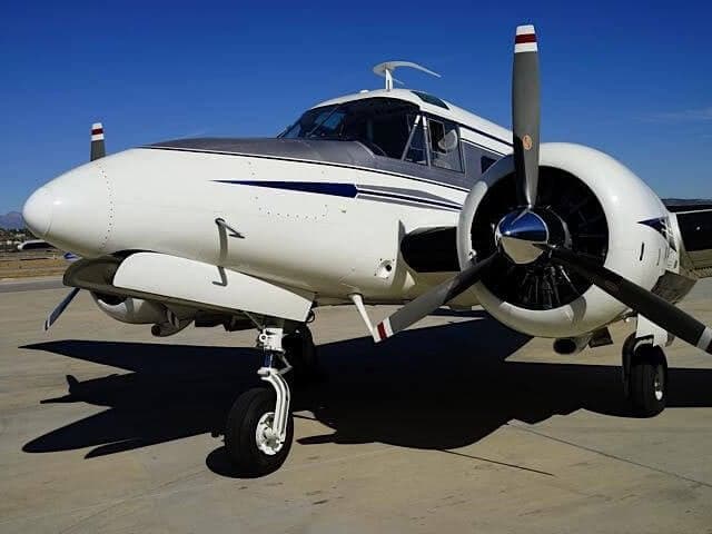 A white and silver Beechcraft King Air with blue stripes parked on an airfield under a clear blue sky.