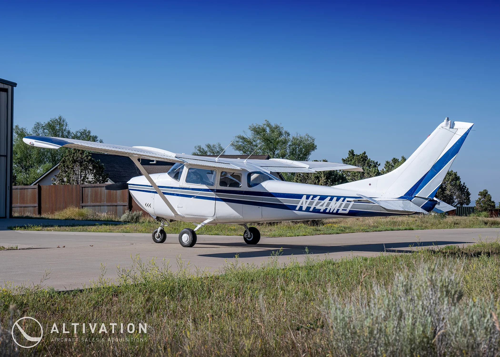 A white Cessna 182 with blue and grey stripes, registration N74MD, parked on a tarmac with trees and a building in the background.