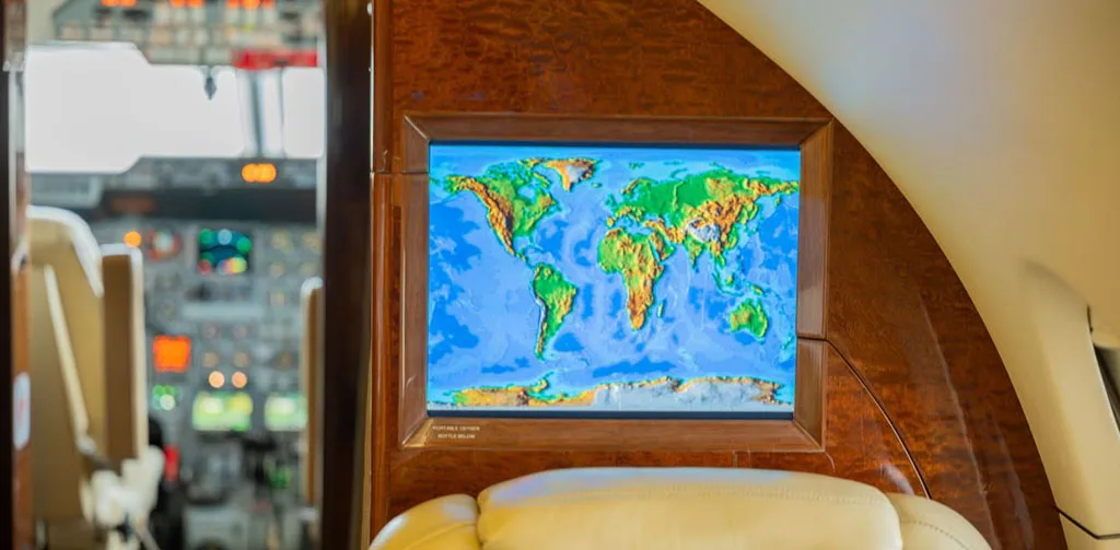 A close-up of a screen displaying a colorful topographical world map, mounted on wood paneling in the private jet cabin.