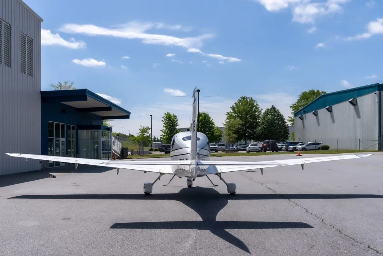 A rear view of a silver and white Cirrus SR22T aircraft parked on an airport tarmac with buildings and trees in the background.