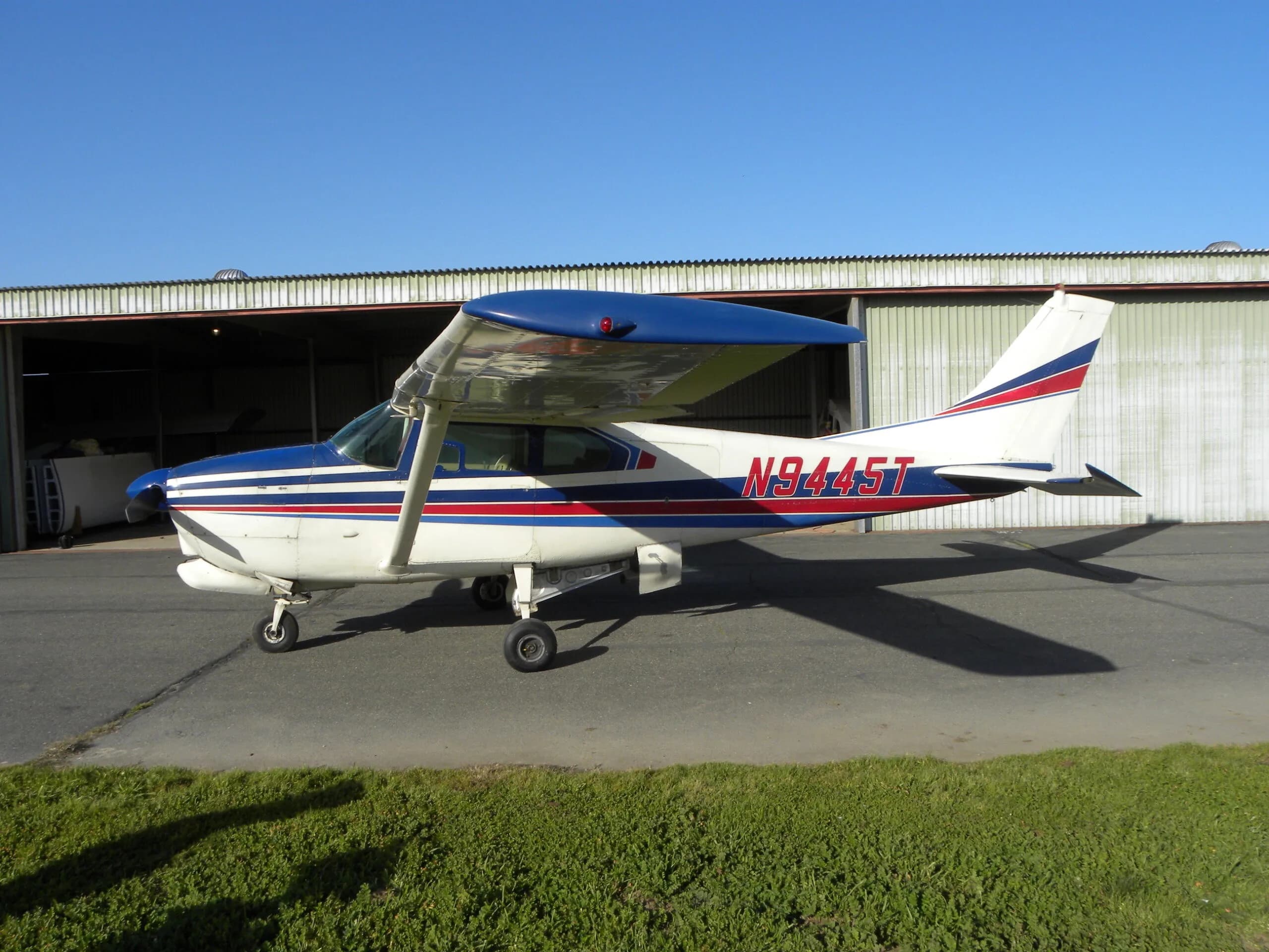 A side profile of a white Cessna 182 with blue and red stripes, registration N9445T, parked on a tarmac.