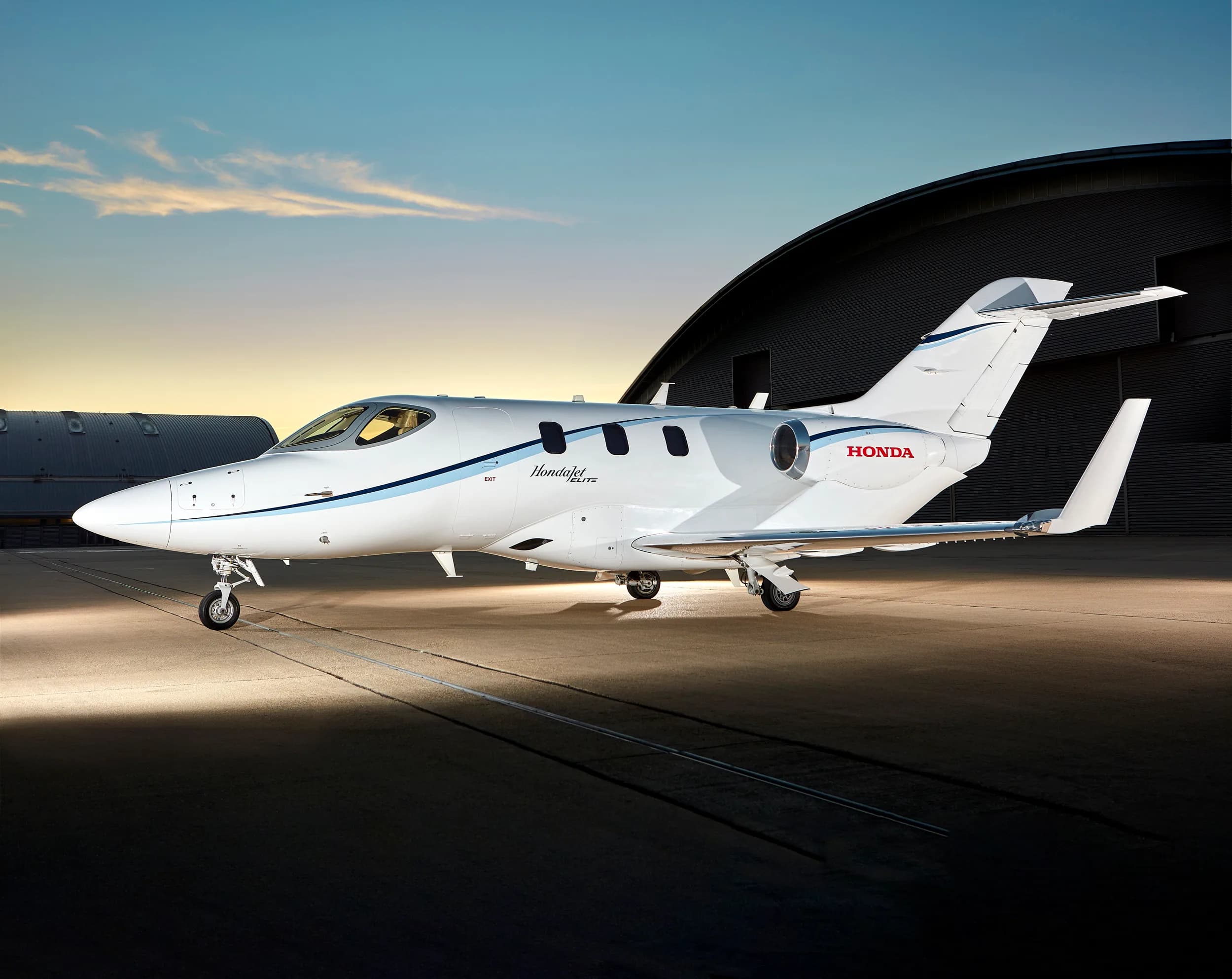 A white HondaJet Elite with blue stripes is parked on a tarmac in front of a hangar at sunset.