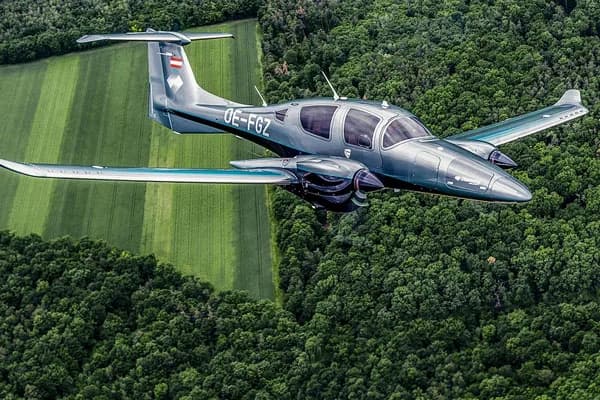 A grey Diamond DA62 twin-engine aircraft flying over green fields and forests.