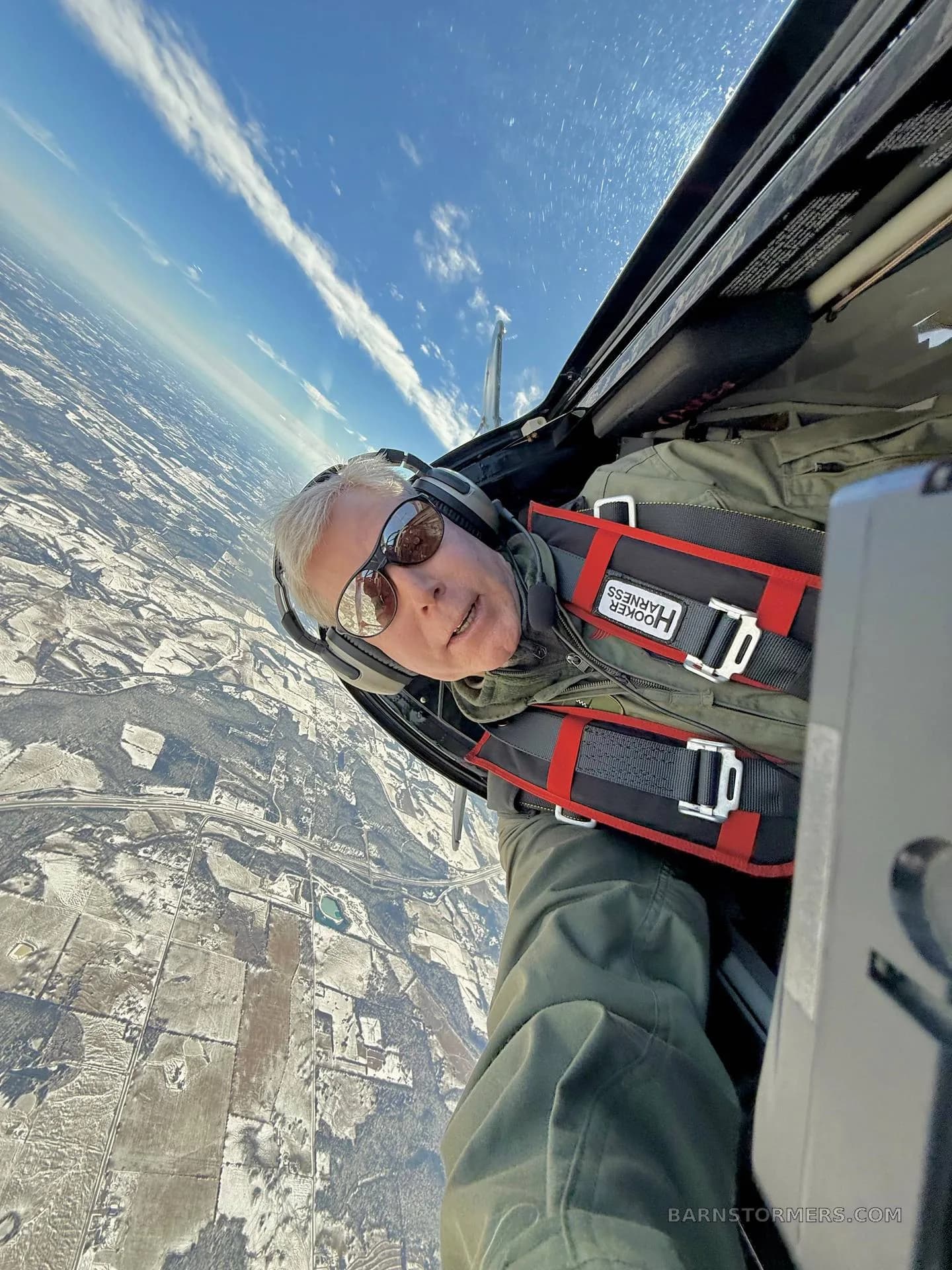 A pilot in a Pitts biplane cockpit, wearing a harness and sunglasses, looking at the camera while flying over a snow-covered landscape.