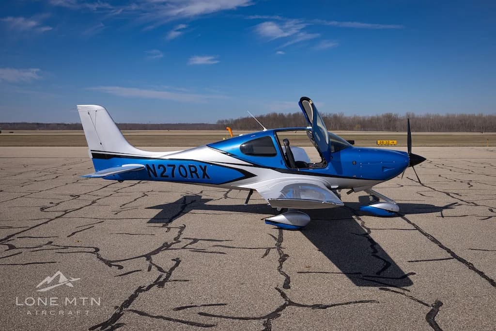 A side view of the blue and silver Cirrus SR20 aircraft, registration N270RX, with its doors open on an airport tarmac.
