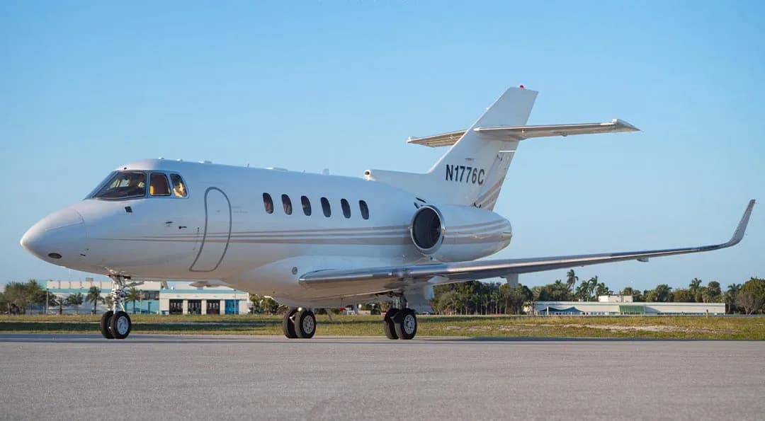 A white Hawker 800 series business jet with a light grey stripe is parked on an asphalt tarmac under a clear blue sky.