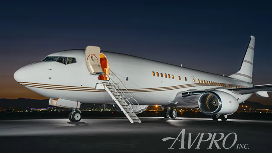 A close-up, front-side view of a white Boeing 737 business jet with brown stripes on the tarmac at night, with its main door open and stairs extended.