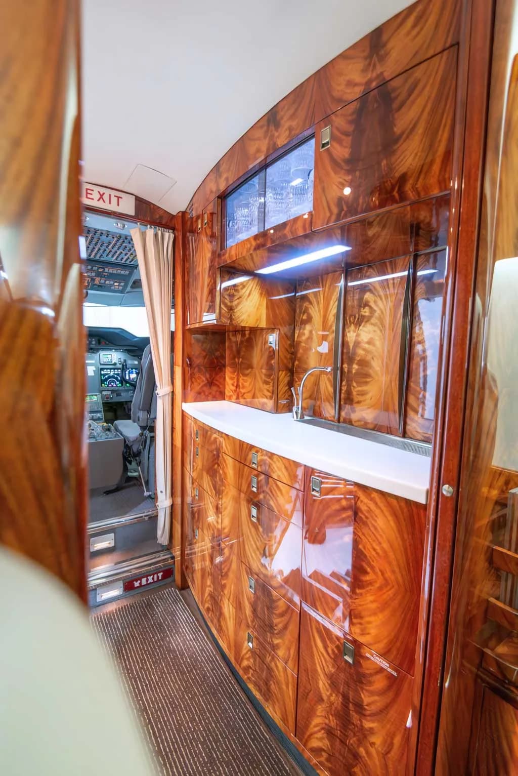 The private jet's galley area with polished wood cabinetry, a sink, and a view towards the cockpit.