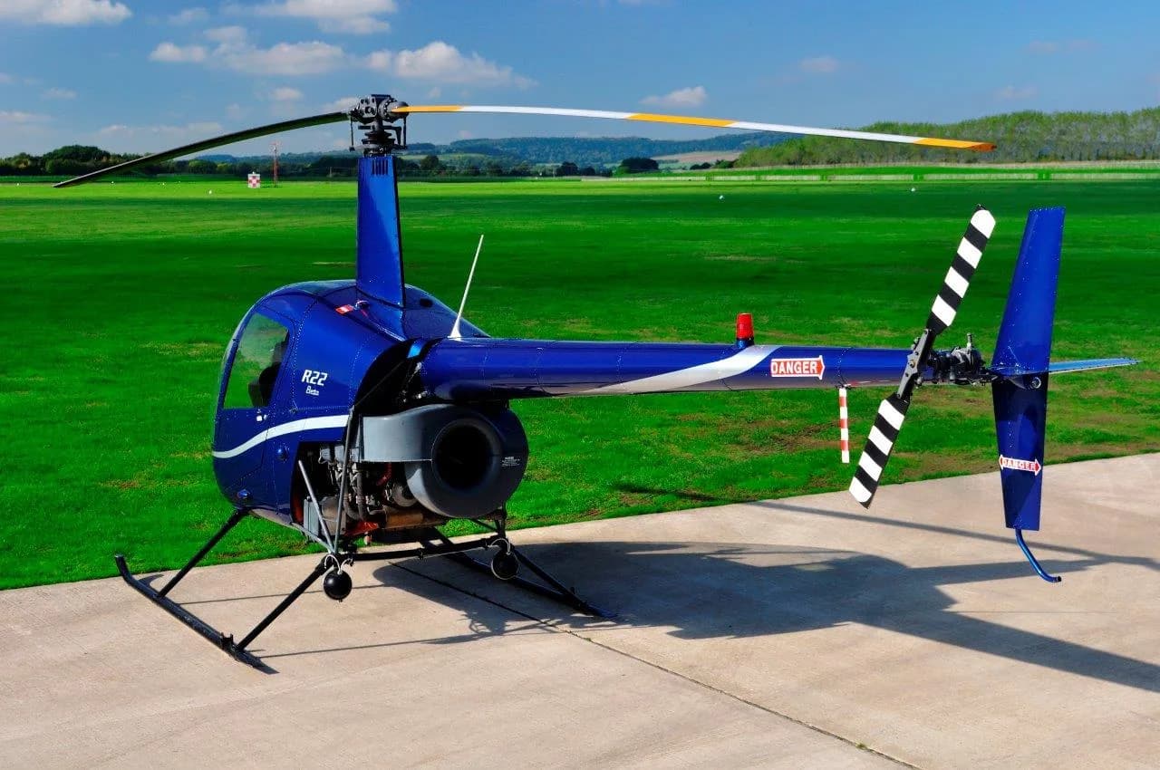 A blue Robinson R22 helicopter with white stripes parked on a tarmac next to a green field under a blue sky.