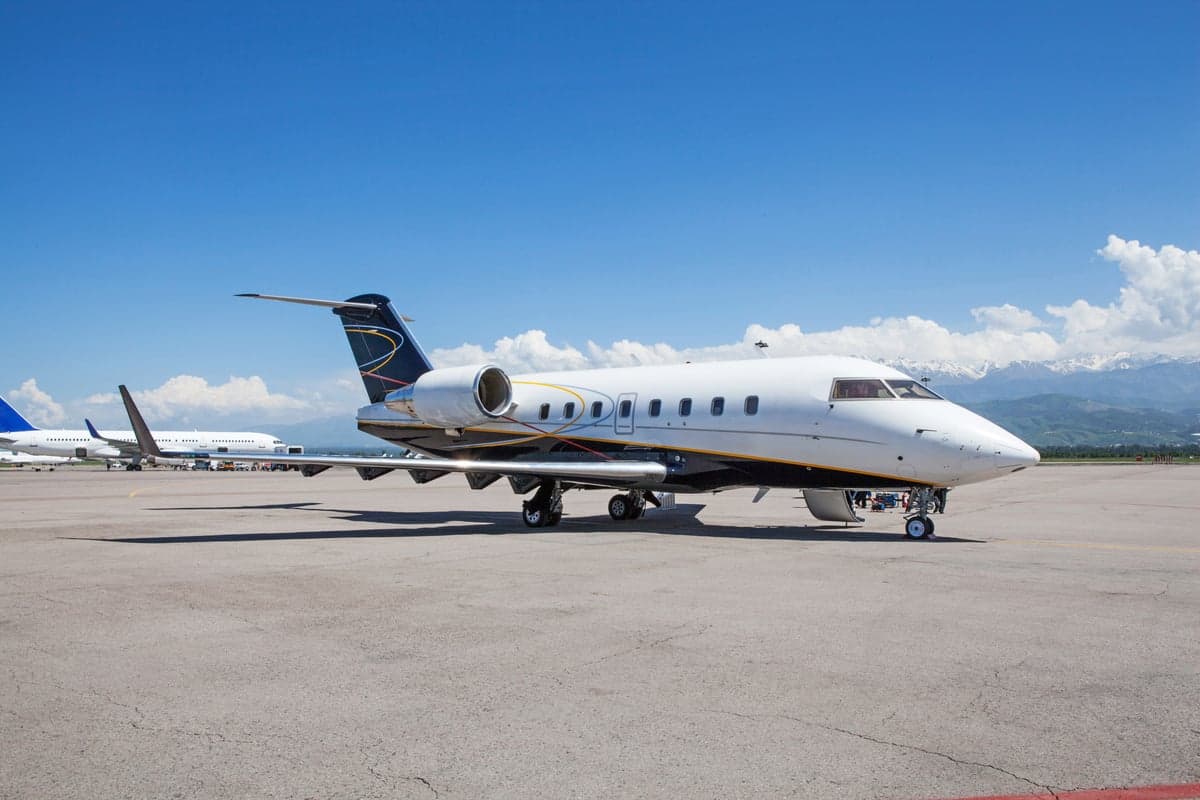 A private jet with a white and blue paint scheme is parked on the tarmac with mountains in the background.