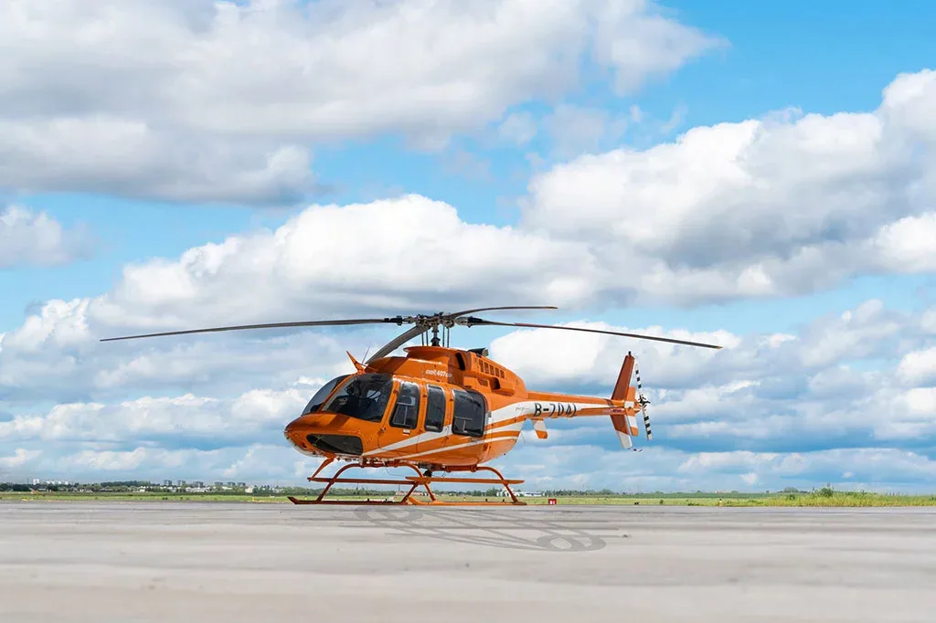 An orange Bell 407 helicopter with white stripes parked on an airfield under a cloudy sky.