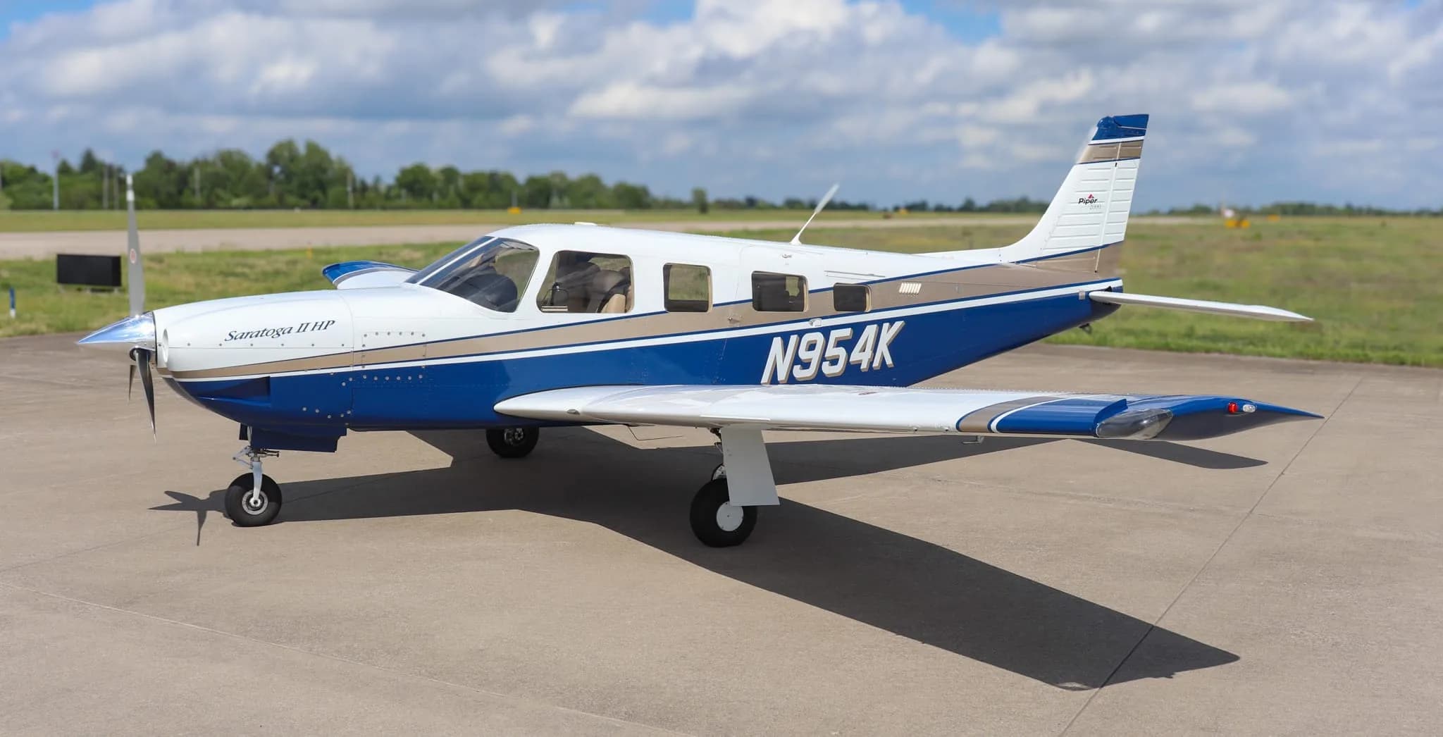 A three-quarter front view of the Piper Saratoga II HP aircraft, N954K, parked on an airfield with trees in the distance.