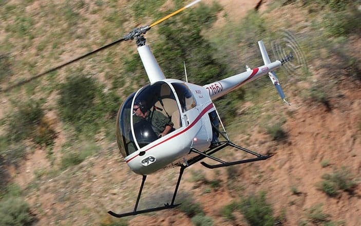 A white Robinson R22 helicopter flying over a desert landscape.