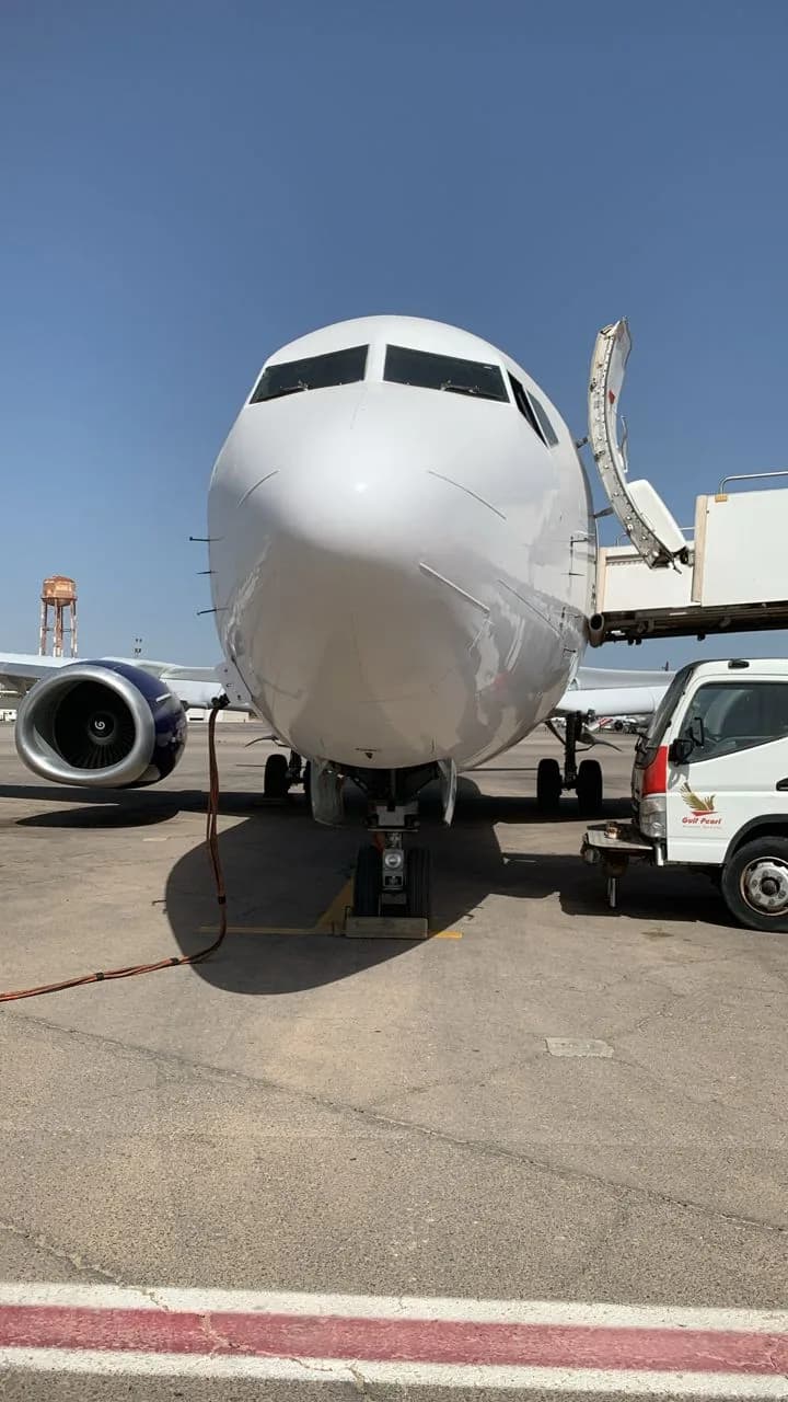 A large white commercial jet, possibly a Boeing 737, is parked on the tarmac with its front door open and a service vehicle nearby.