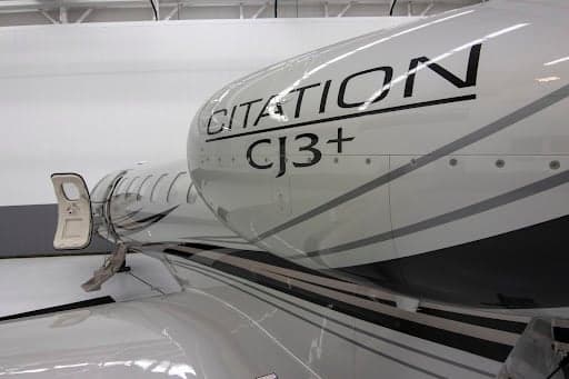 A close-up view of the engine nacelle and fuselage of a silver Cessna Citation CJ3+ aircraft, showing the "CITATION CJ3+" text and an open passenger door.