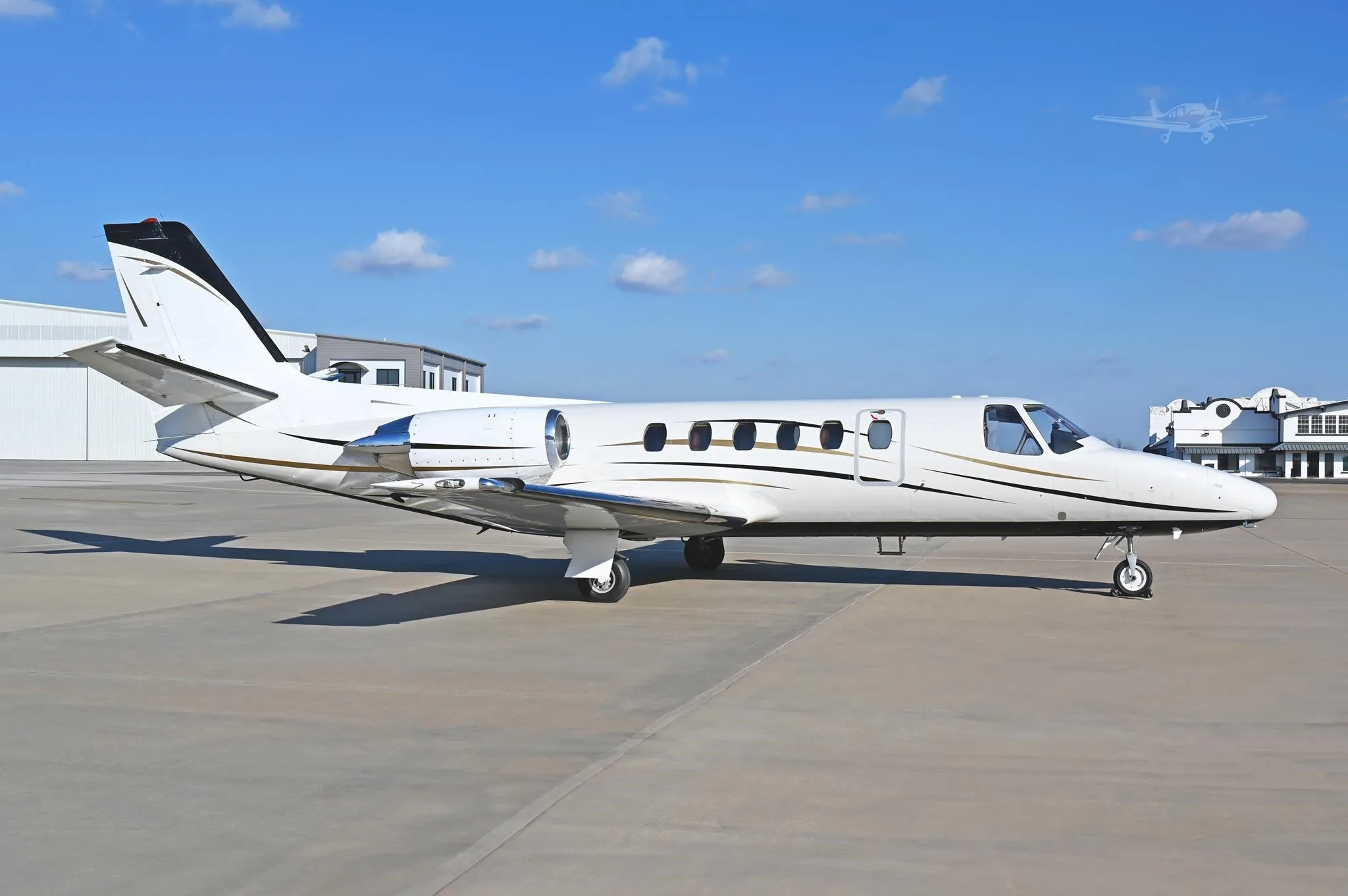 A white business jet with black and gold accent stripes is parked on an airport tarmac under a clear blue sky.