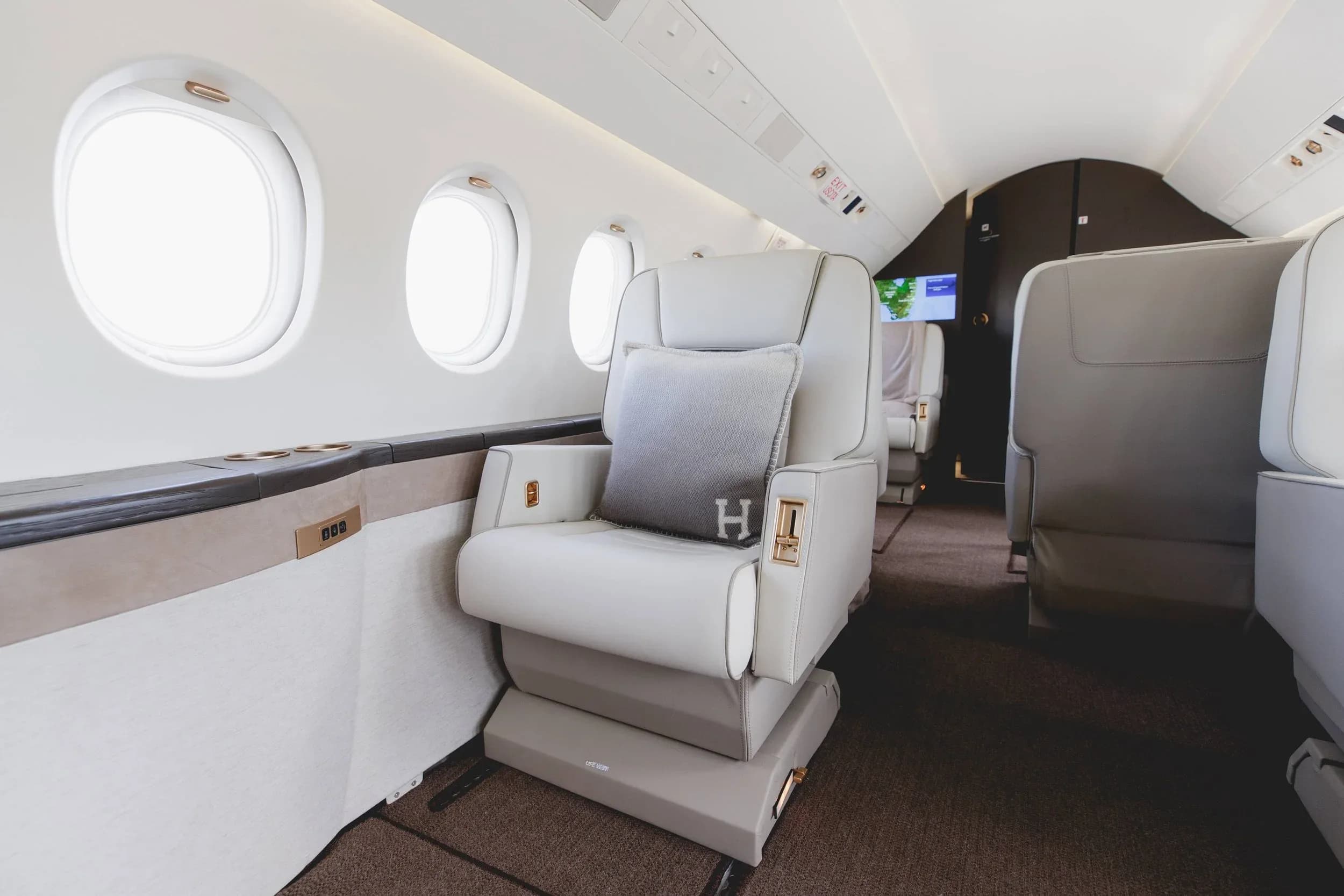 A close-up view of a plush light-colored leather seat with an "H" monogrammed pillow inside a private jet cabin.