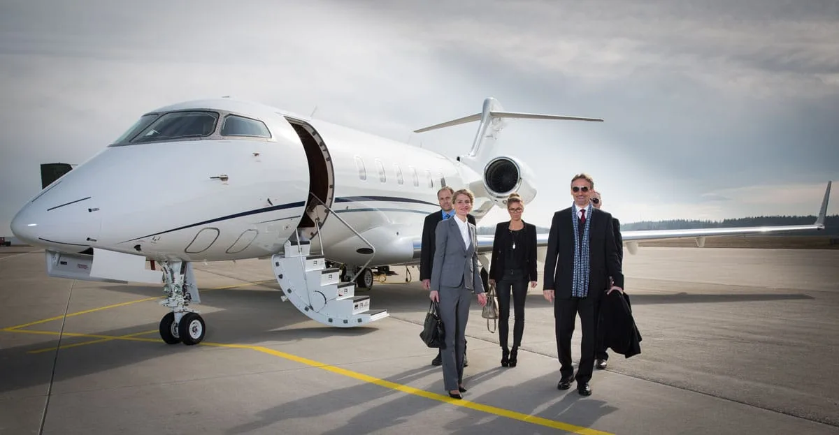 A group of business people walk away from a white Bombardier Challenger private jet with its door open and stairs extended on an airfield.