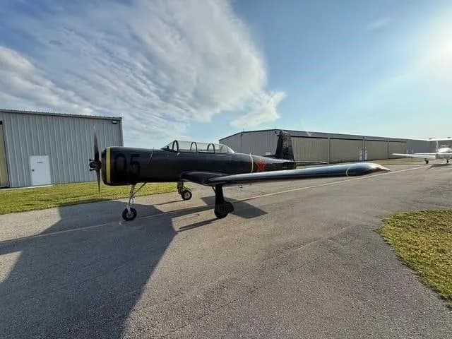 A dark gray Yak-52 aircraft with red stars and yellow accents parked on an asphalt ramp under a partly cloudy sky.