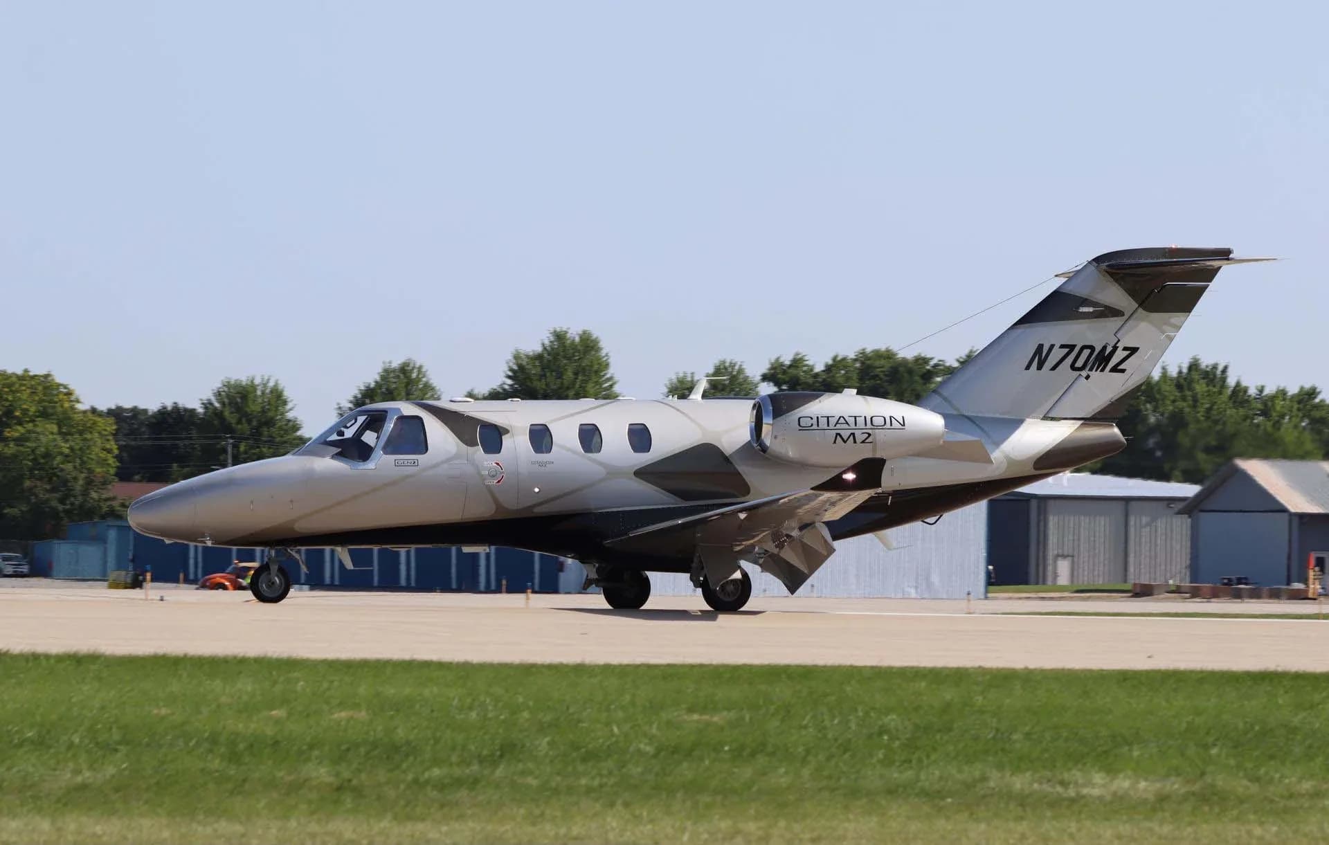 A side view of a silver and black Cessna Citation M2 jet (N70MZ) on a taxiway with green grass and buildings in the background.