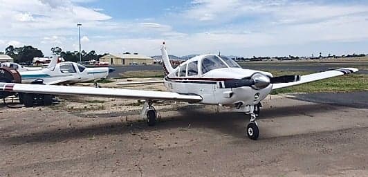 A white single-engine propeller aircraft with dark stripes is parked on an airfield, with another aircraft partially visible in the background.
