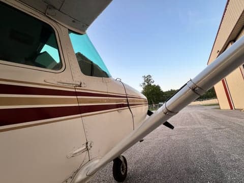 A close-up view of the cockpit side and wing strut of the Cessna 172, N97055, with a hangar in the background.