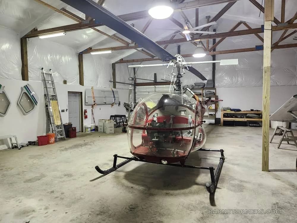 A red and white helicopter with a clear bubble canopy is parked in a hangar with a small plane in the background.