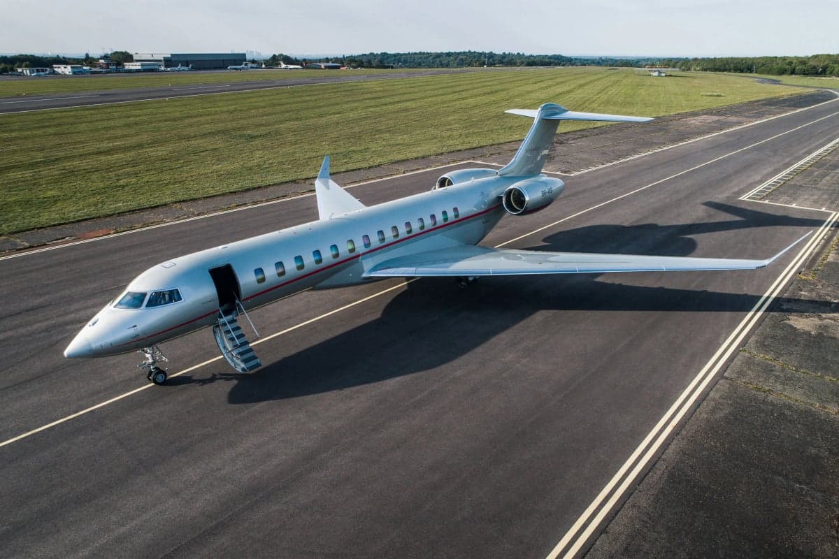 A silver Bombardier Global 6000 business jet with a red stripe on a runway, with its main door open and stairs extended.