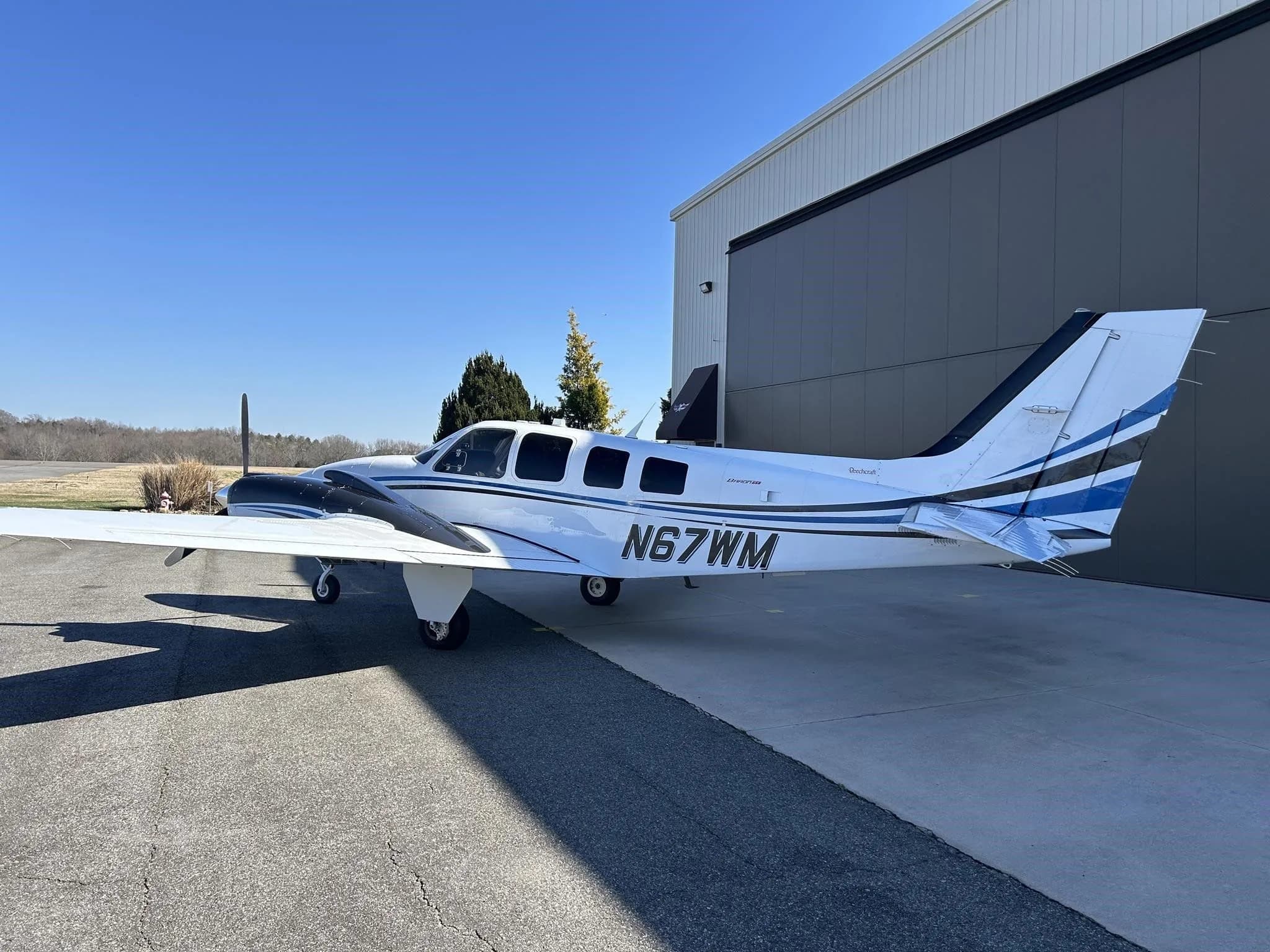 A white and blue Beechcraft Baron N67WM parked on an tarmac outside a hangar, viewed from the side.