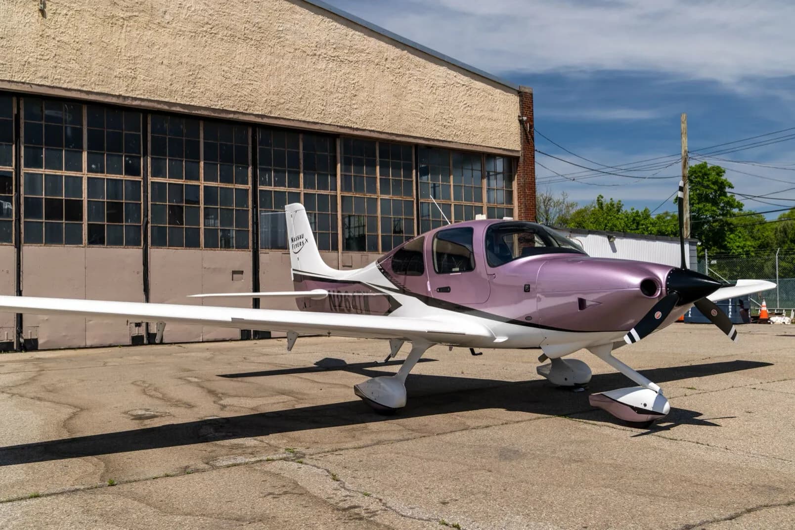 A Cirrus SR22 aircraft with a metallic lavender and white paint scheme, registration N264TM, parked on an airfield in front of a hangar.
