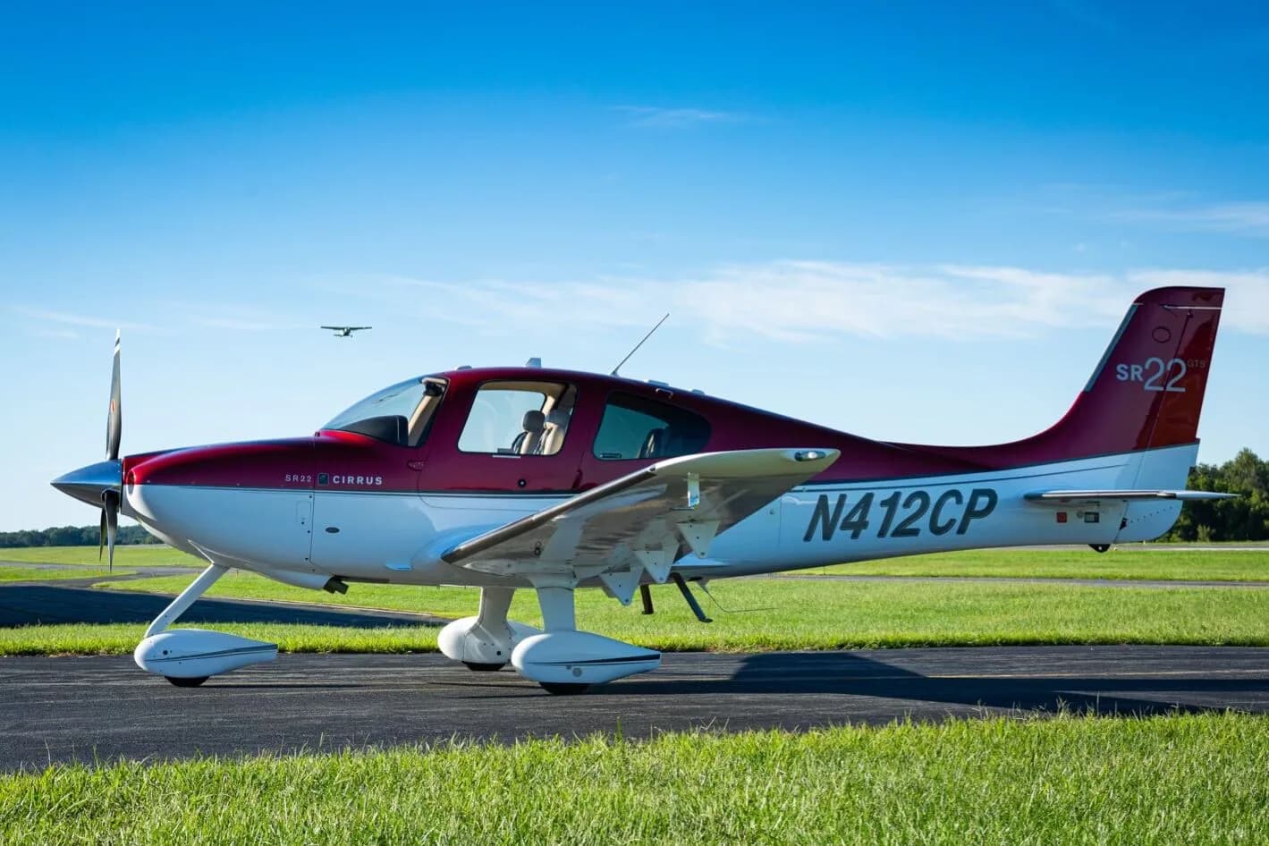 A red and white Cirrus SR22 aircraft (N412CP) parked on an airfield with another aircraft flying in the distance.