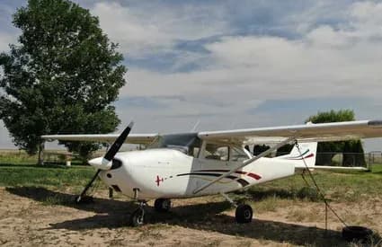 A white single-engine propeller aircraft with dark and red stripes is parked on a dirt field with a large tree in the background.