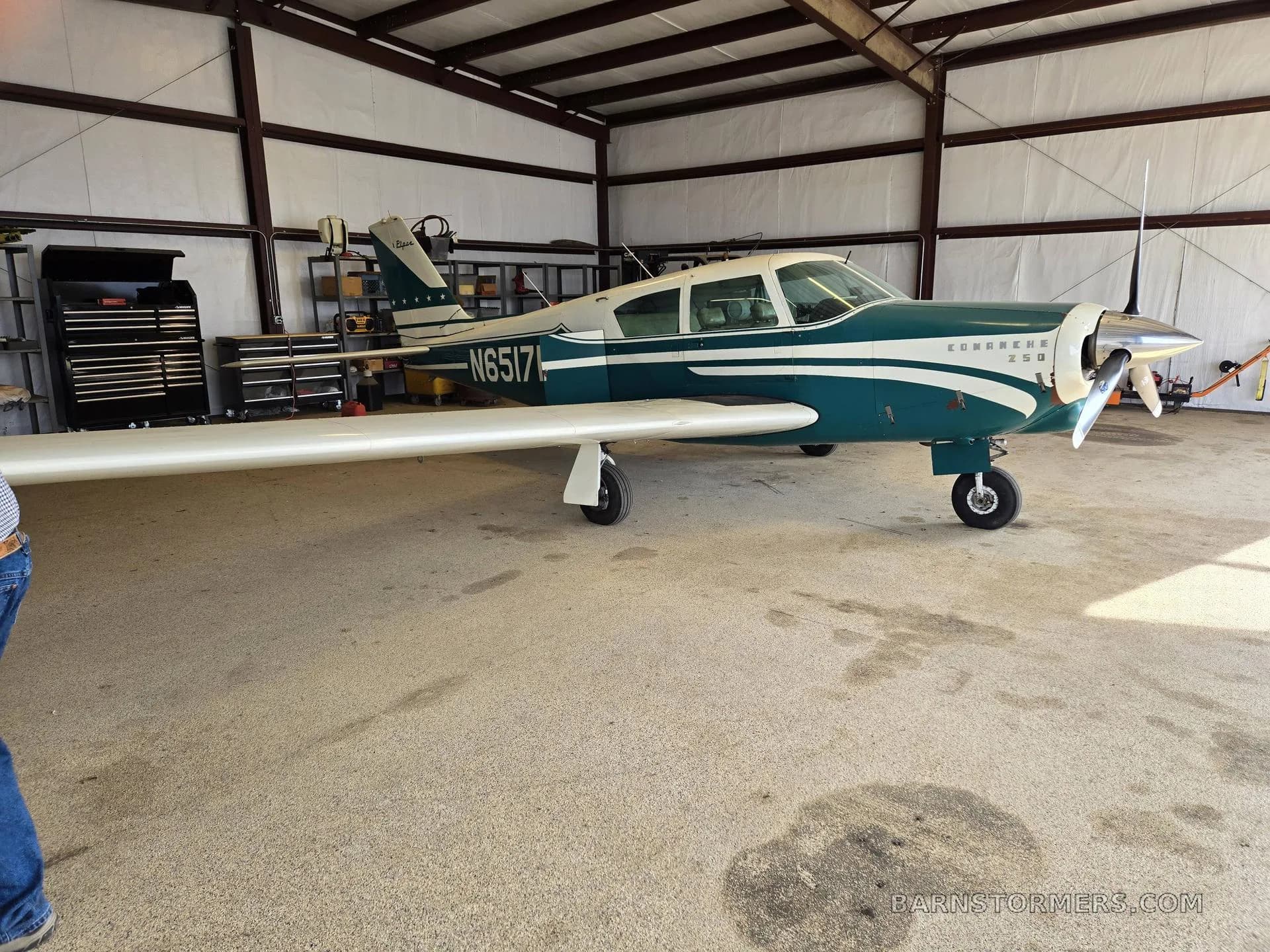 A side view of a white and teal Piper Comanche 250 aircraft with registration N6517P, parked inside a hangar.