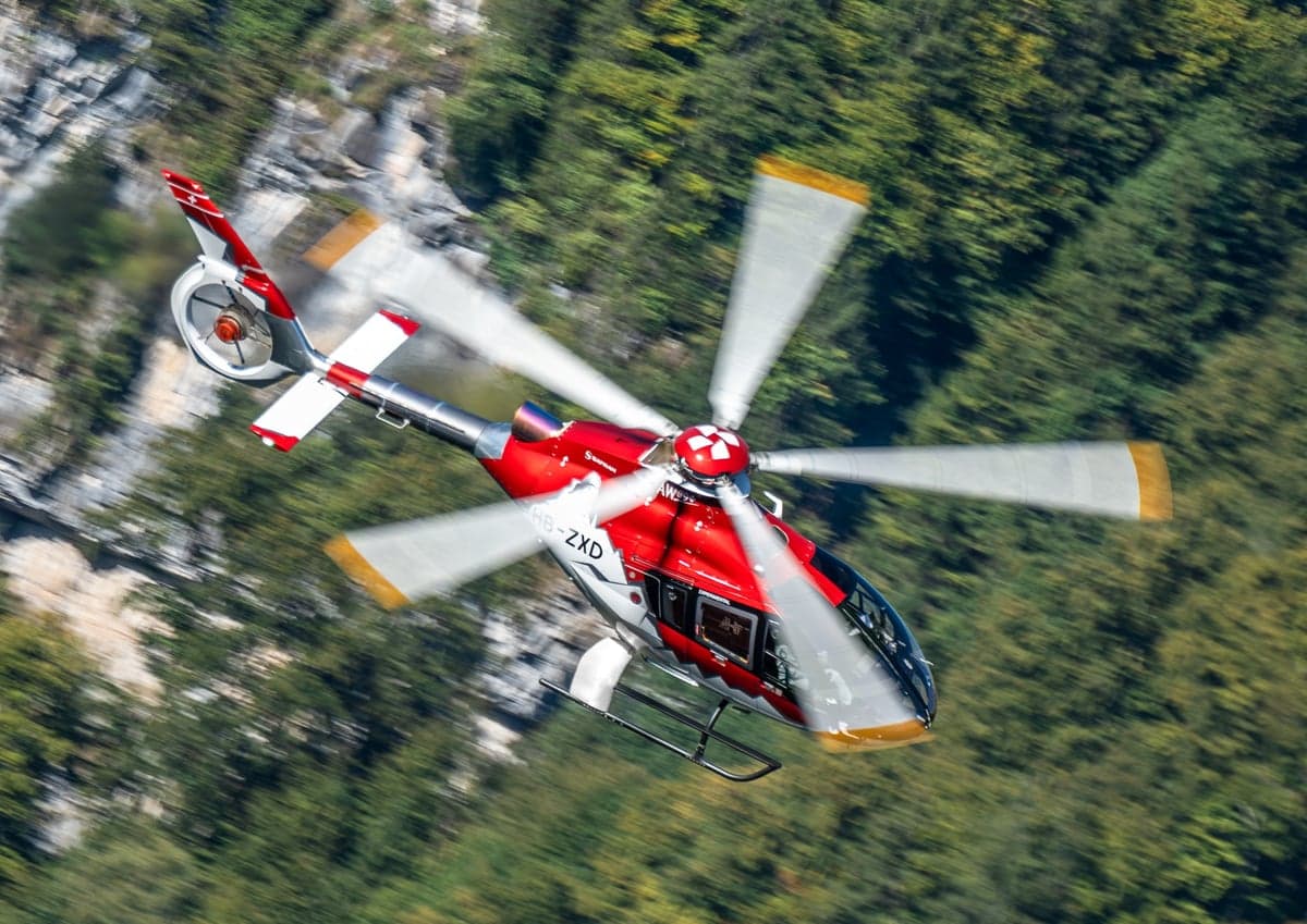 A red and white Airbus H135 helicopter in flight near mountains.