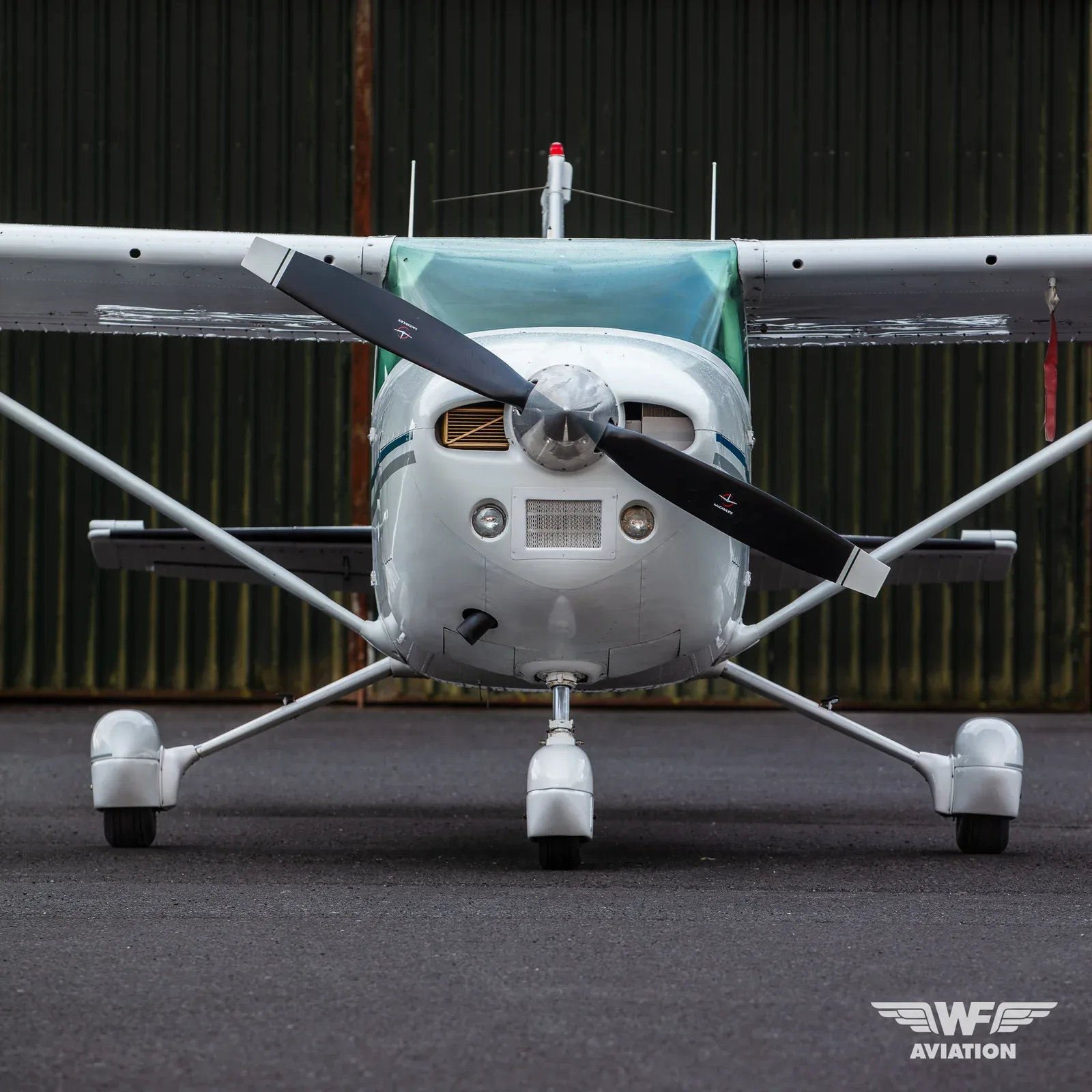 Front view of a white single-engine propeller aircraft with a blue stripe, parked on asphalt in front of a dark hangar.