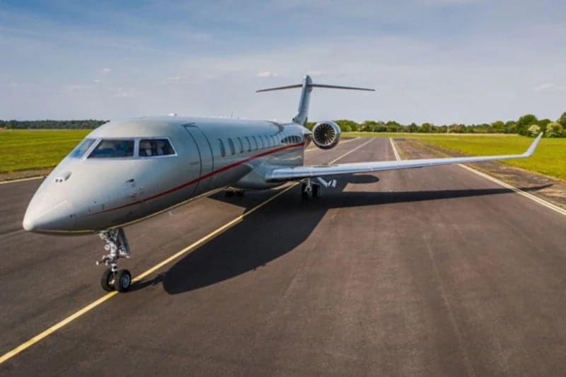 A silver Bombardier Global 6000 business jet with a red stripe parked on a runway under a clear sky.