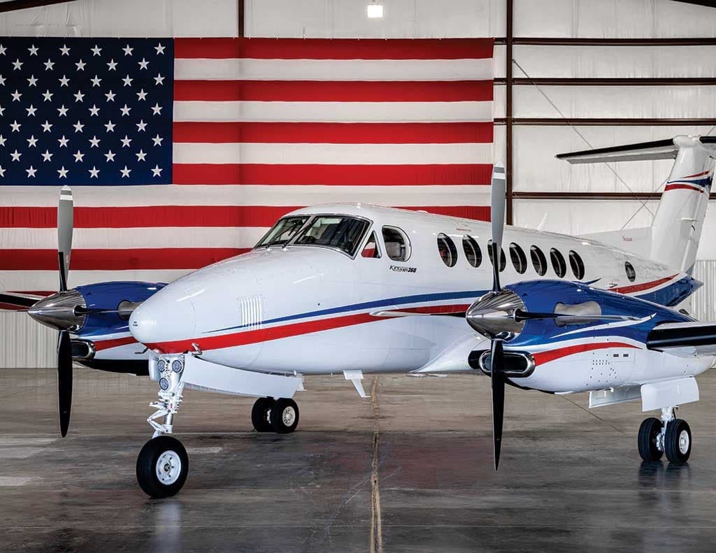 A white Beechcraft King Air 360 with blue and red stripes parked in a hangar in front of a large American flag.