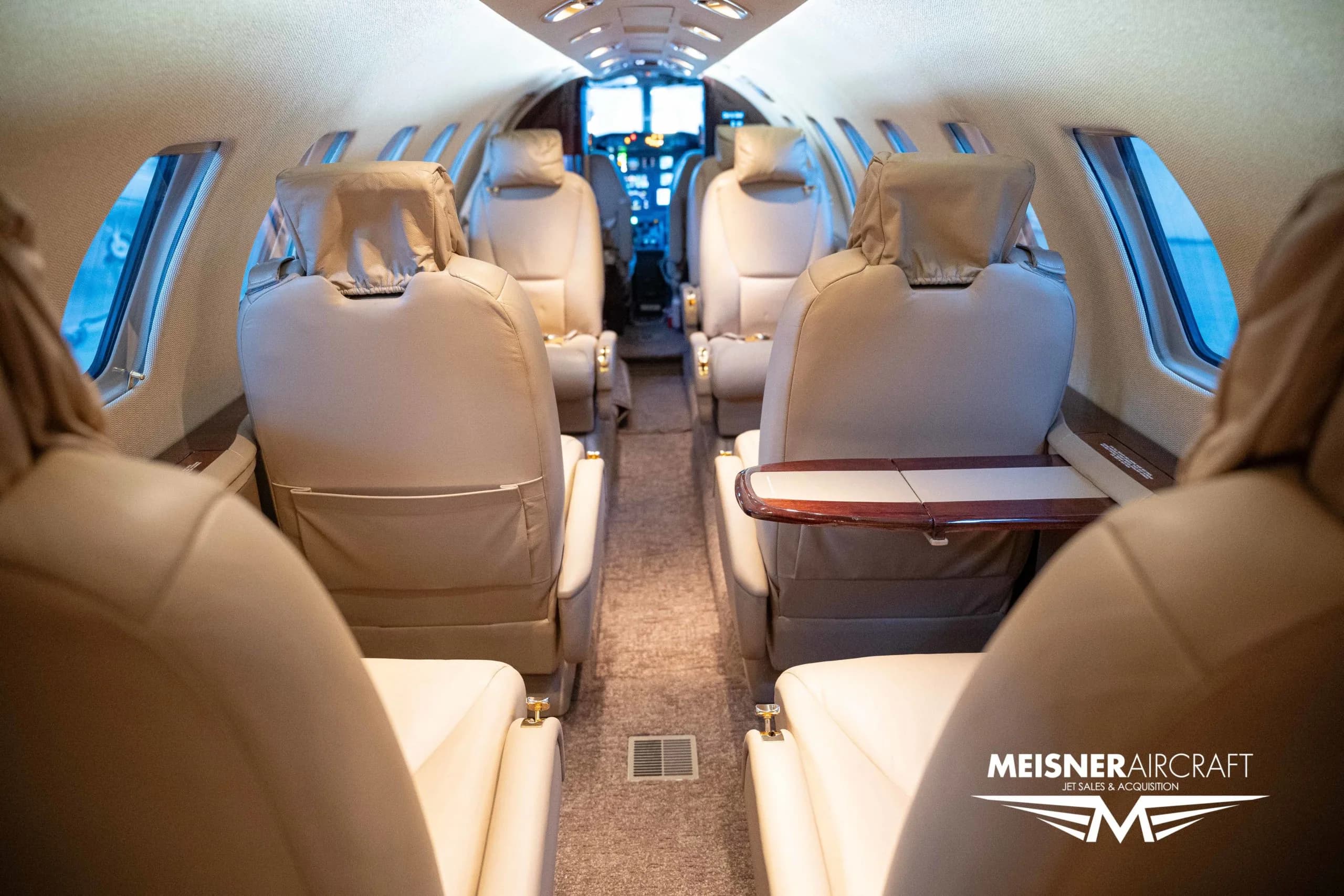 The interior cabin of a private jet, featuring beige leather seats and a fold-out table, looking towards the cockpit.