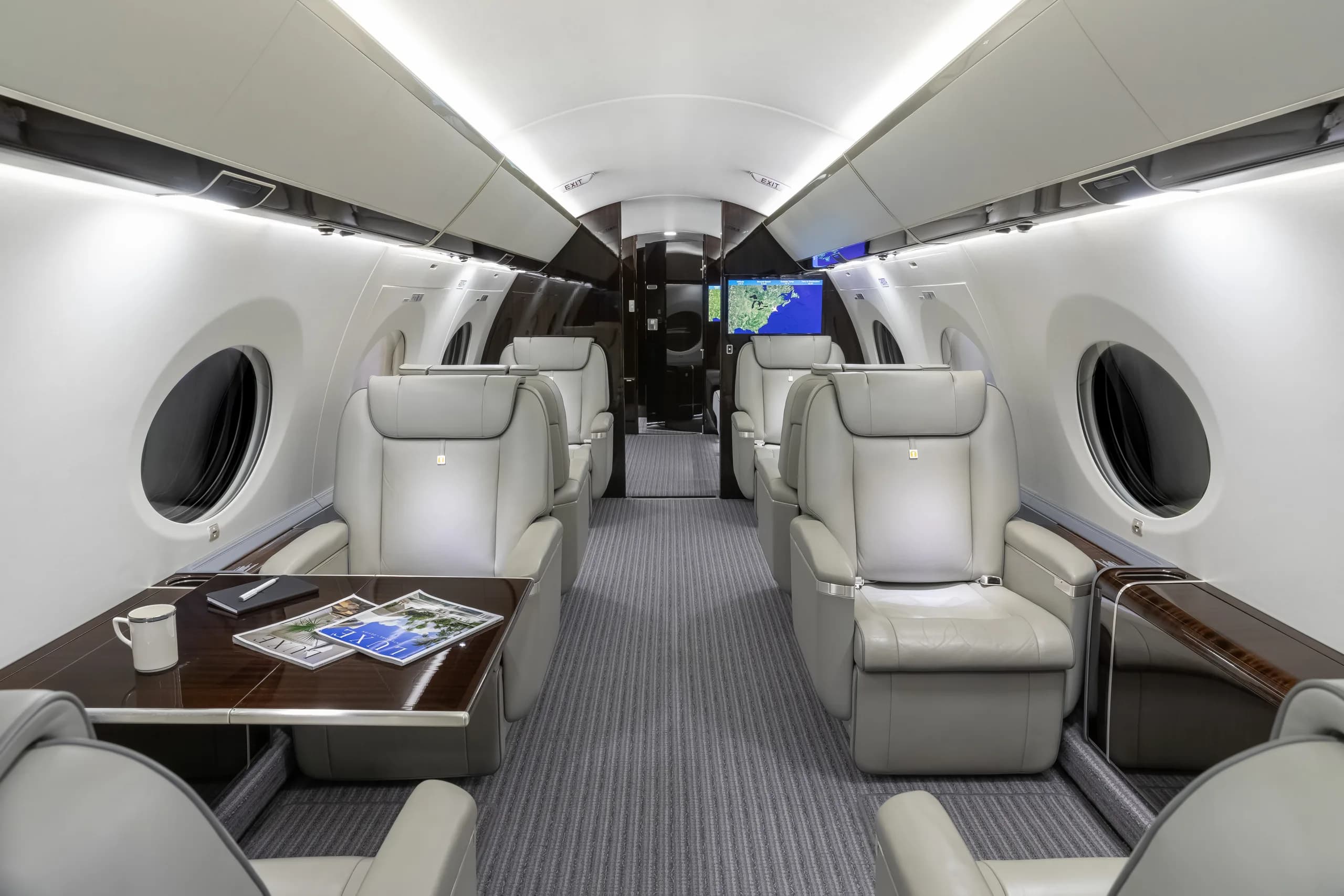 The luxurious cabin interior of a private jet, featuring light grey leather seats, a dark wood table, and a map displayed on a screen at the rear.