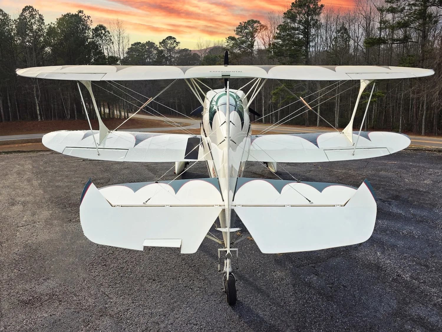 A rear view of the white biplane with green accents, parked on a gravel surface at sunset.