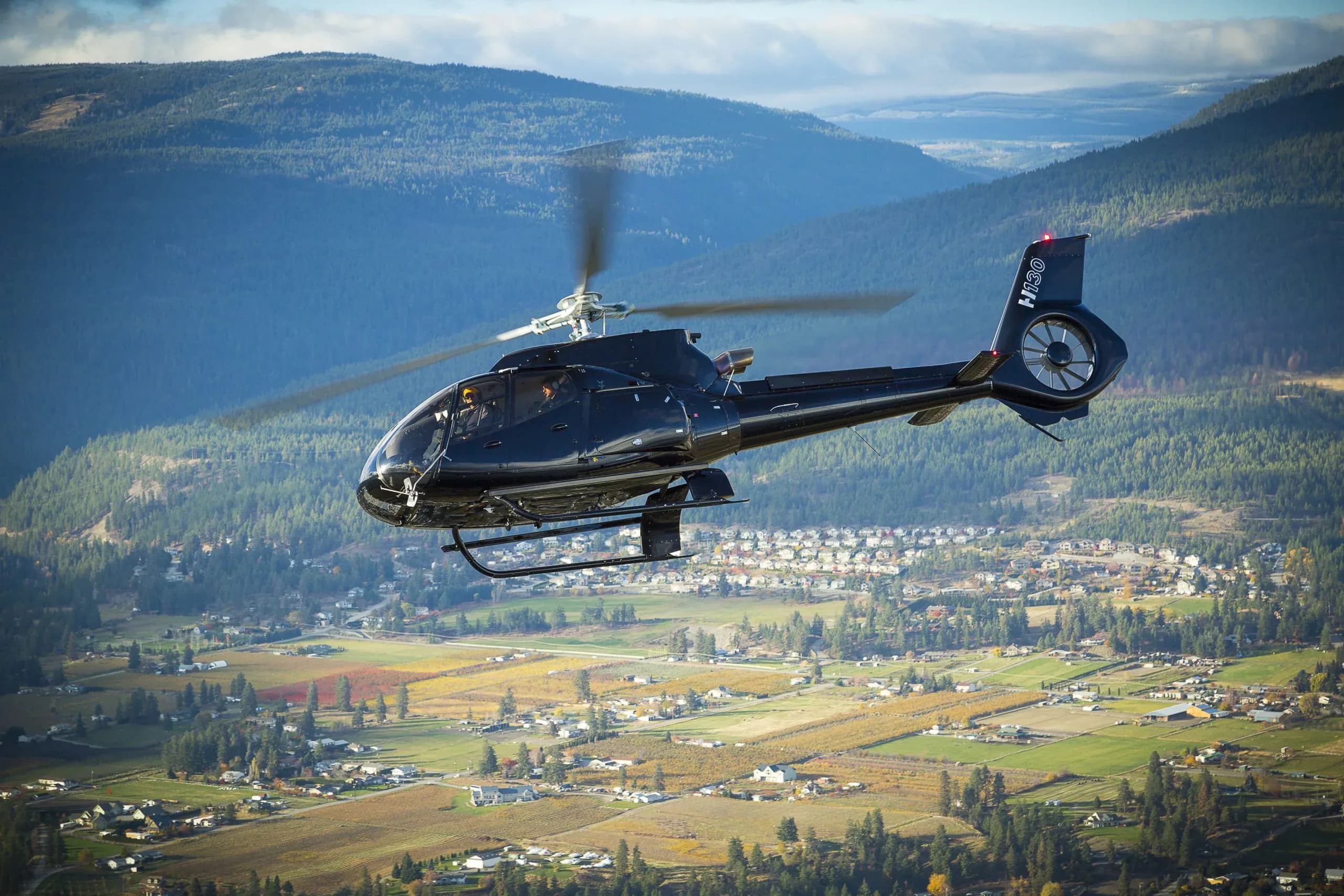 A black Airbus H130 helicopter flies over a scenic valley with a town and autumn fields, backed by forested mountains.