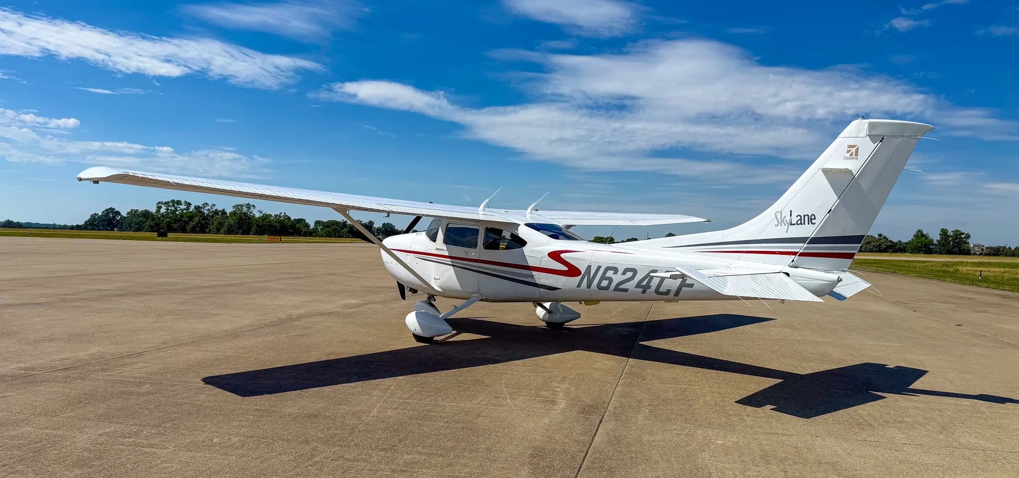 A white Cessna 182 Skylane with red and gray stripes, registration N624CF, parked on an asphalt ramp under a bright blue sky.
