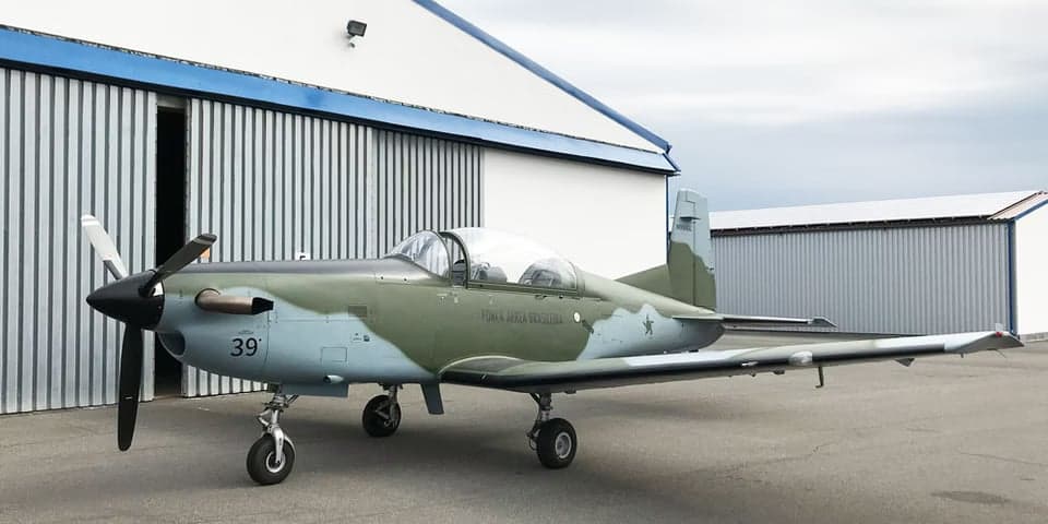 A Tucano aircraft with a camouflage paint scheme parked in front of a hangar.