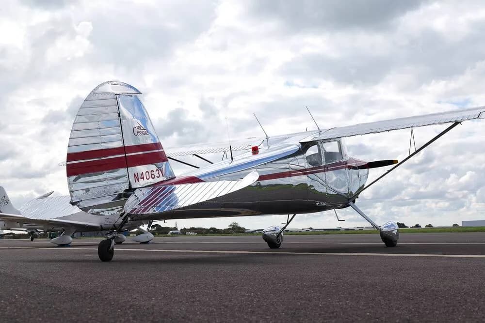 A polished silver Cessna 170B with red stripes on the tail and fuselage, parked on an asphalt tarmac under a cloudy sky.